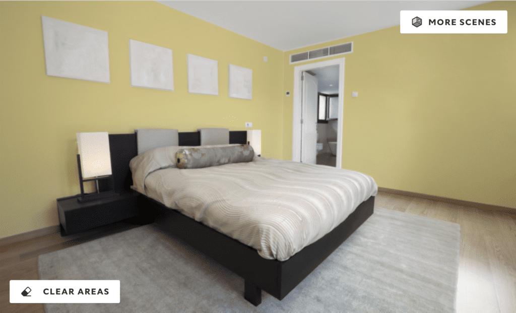 A modern bedroom with vibrant yellow walls, neutral bedding, and minimalist decor. The room features a low-profile bed, black side tables with lamps, and blank picture frames above the bed, with natural light coming from an adjoining bathroom.