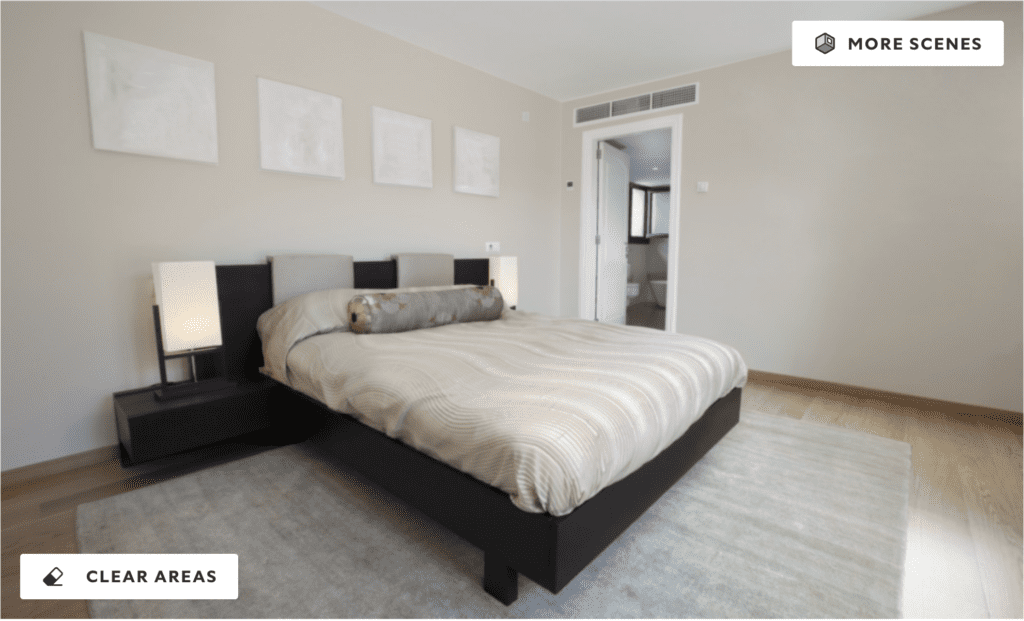 A minimalist bedroom with soft ivory walls, neutral bedding, and modern furnishings. The bed is centered between two black side tables with simple lamps, and the wall above features three blank picture frames. The space is well-lit with natural and ambient lighting.