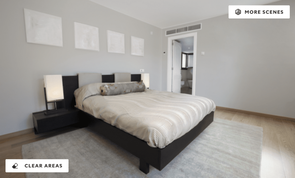 A modern bedroom with soft gray walls, minimalist decor, and neutral bedding. The room features a sleek low-profile bed, black side tables with simple lamps, and blank picture frames above the bed, with light wood flooring and natural lighting from an adjoining bathroom