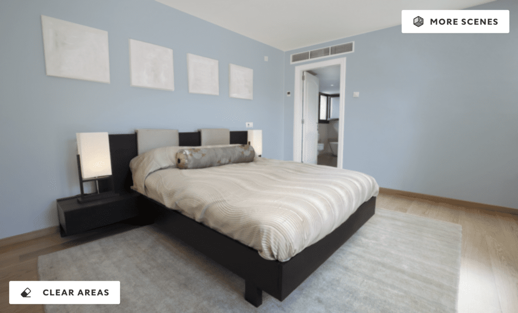 A modern bedroom with soft Moonmist blue walls, neutral bedding, and minimalist decor. The room features a low-profile bed, black side tables with lamps, and blank picture frames above the bed, illuminated by natural light from an adjoining bathroom.