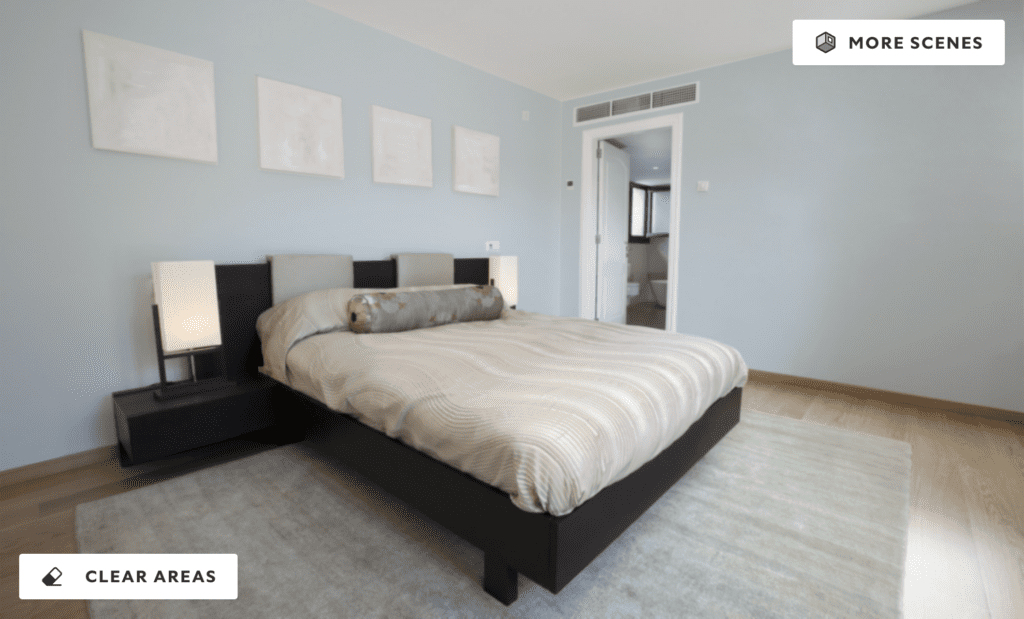 A modern bedroom with soft Sky High blue walls, neutral bedding, and minimalist decor. The room features a low-profile bed, black side tables with lamps, and blank picture frames above the bed, illuminated by natural light from an adjoining bathroom.